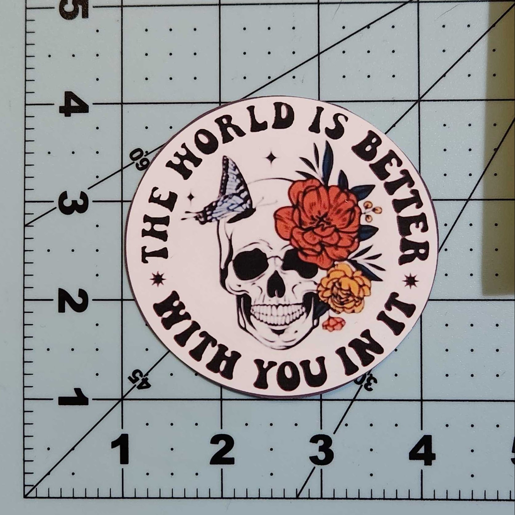 Circular sticker with skull design and text 'The world is better with you in it' on a cutting mat.