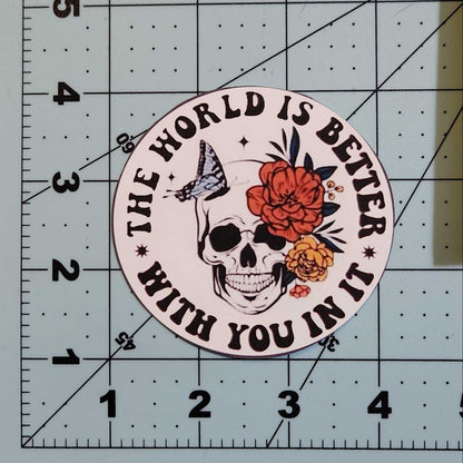 Circular sticker with skull design and text 'The world is better with you in it' on a cutting mat.